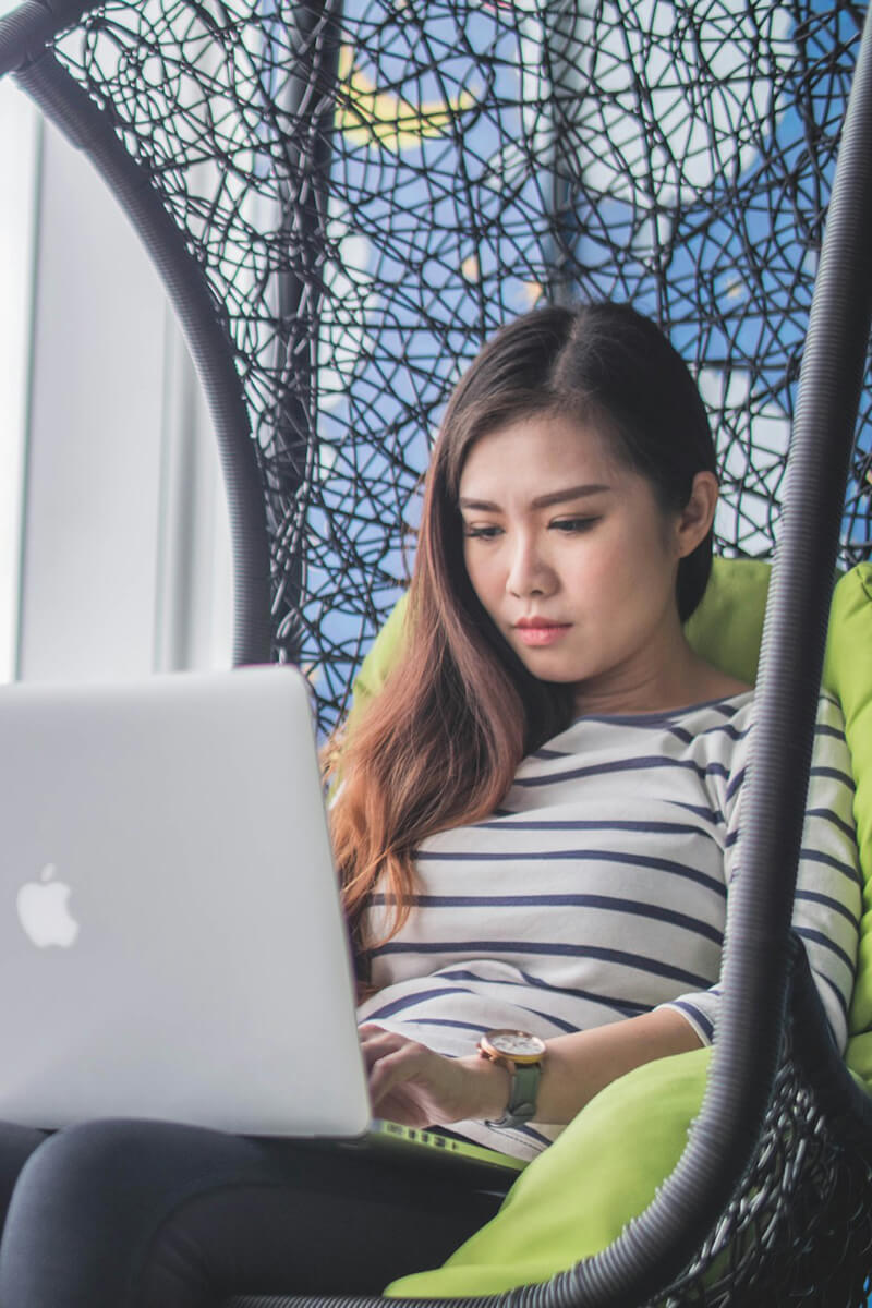 A young Asian woman sitting in a hammock chair on a Macbook laptop.