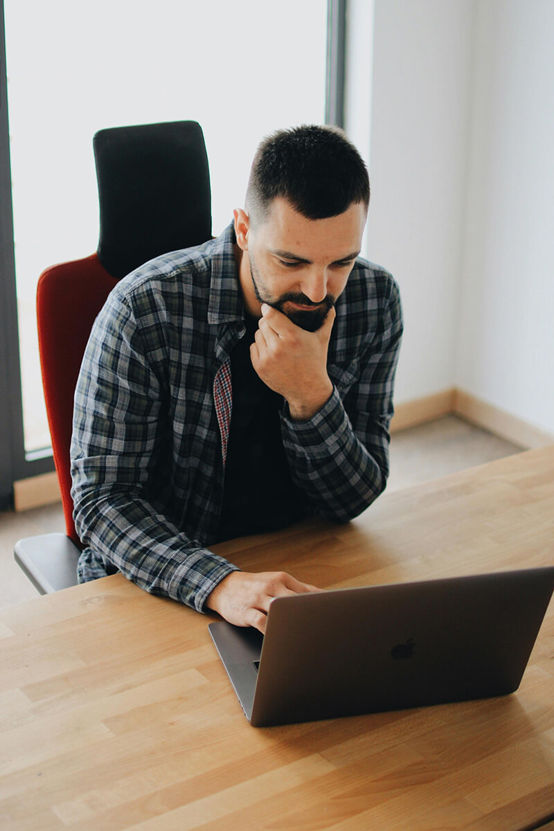 A man sitting at a desk looking thoughtfully at a Macbook laptop screen.