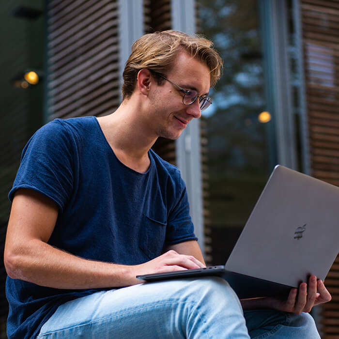 A young man with glasses sitting outside and smiling while looking at his laptop.