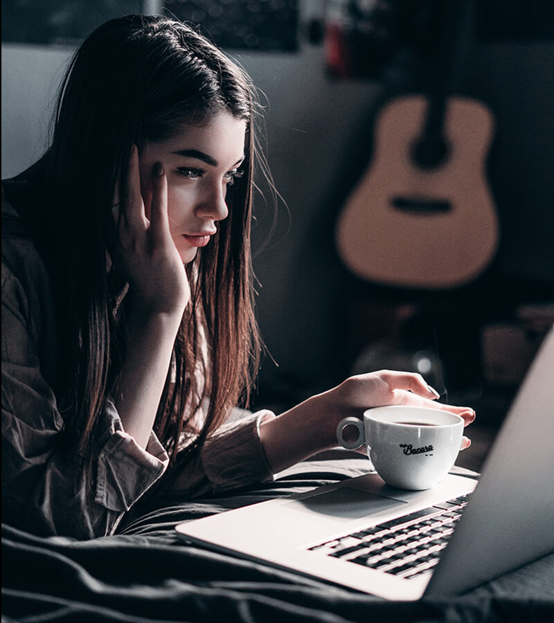 A young woman with a cup of coffee looking intently at a laptop screen.