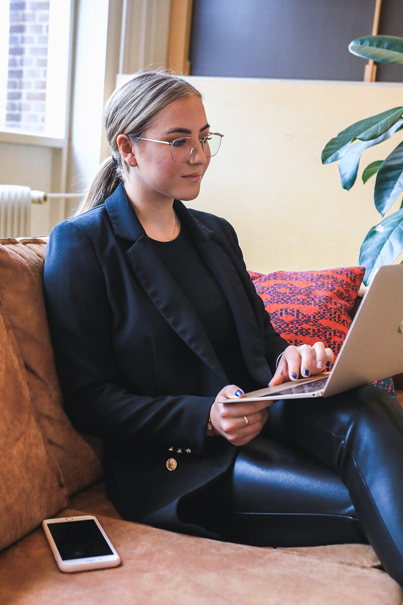 A woman in business attire sitting on a couch with a laptop.